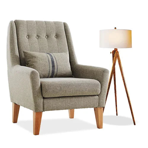 Cozy gray armchair with tufted backrest, wooden legs, and a lumbar pillow paired with a sleek tripod floor lamp, creating a warm, inviting atmosphere.