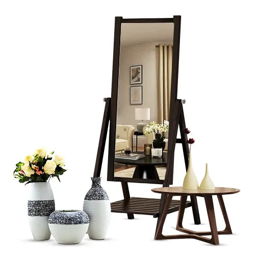 A tall, dark-framed mirror reflects a cozy living room with a sofa and lamp. Vases with flowers and a small wooden table with decor sit nearby.