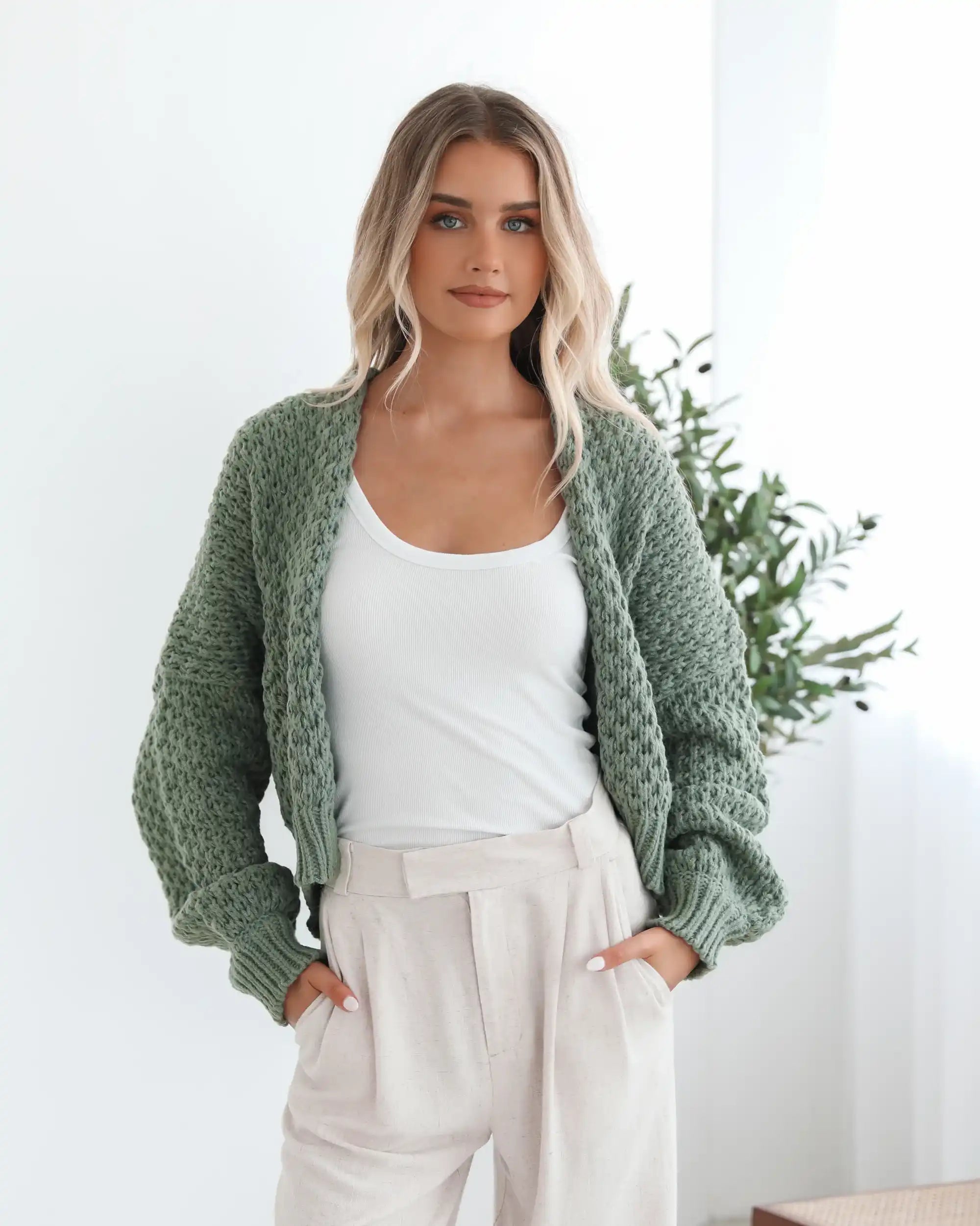 A woman with long blonde hair wears a green knit cardigan over a white tank top and beige pants. She stands confidently with hands in pockets, against a leafy background.