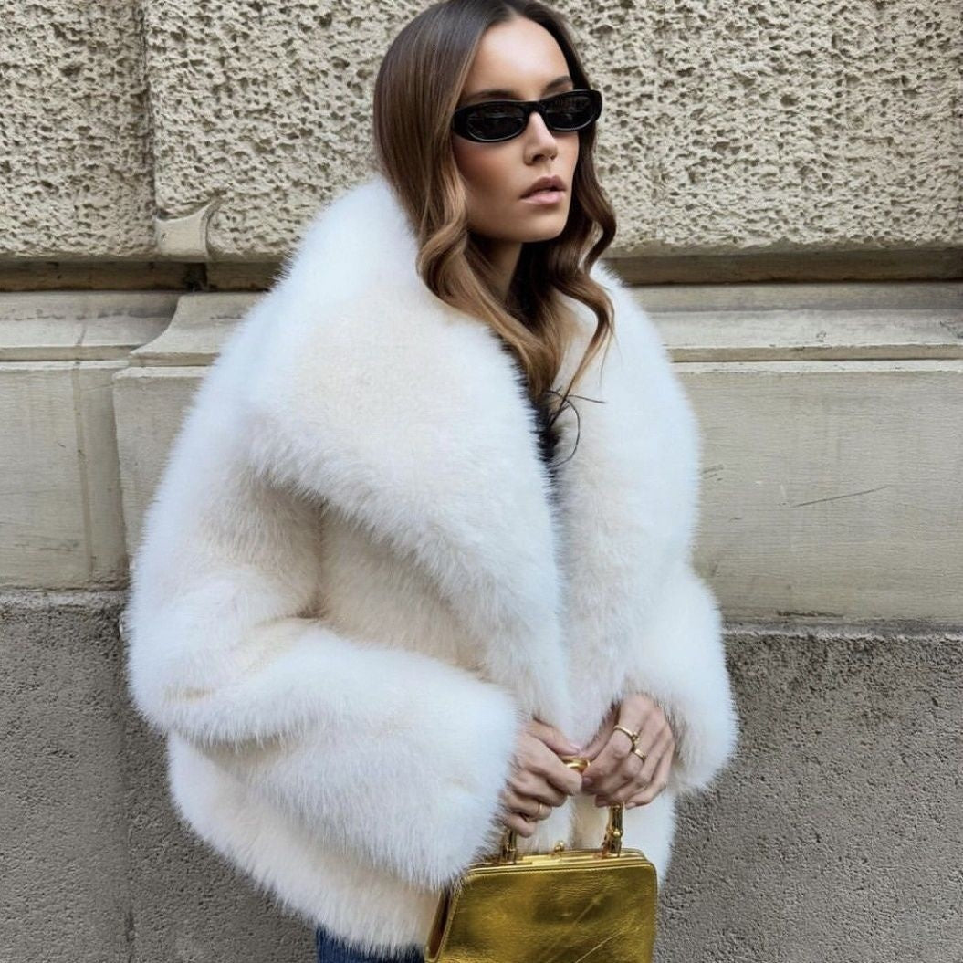 Woman wearing a white fur coat and sunglasses, holding a gold handbag against a stone wall.