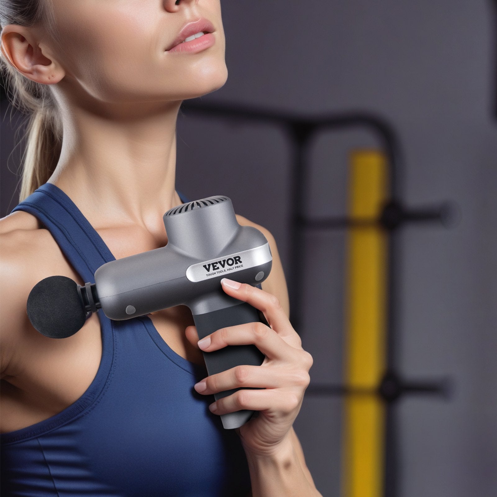 Woman using a VEVOR massage gun on her shoulder with a blurred gym background